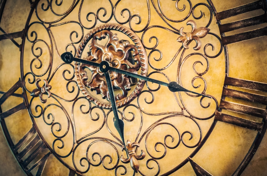 An ornate wall clock featuring intricate iron scrollwork, a central decorative motif, and long, slender hands against a muted background