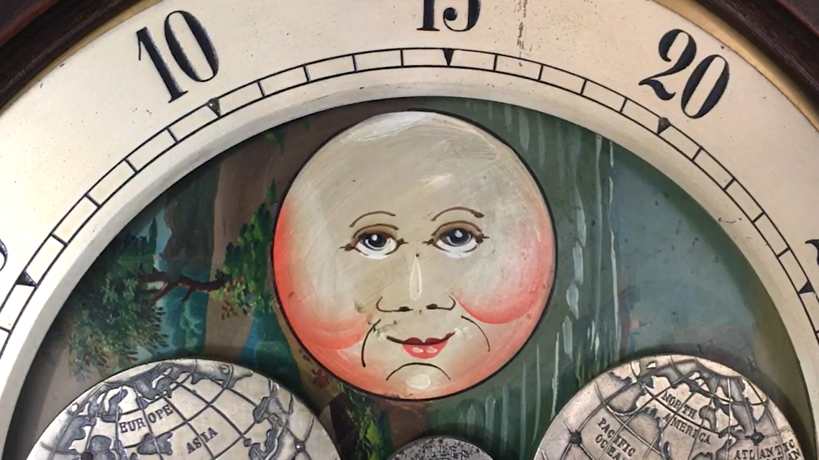 A vintage clock displays a painted moon with a face, with small illustrations of a forest landscape and a map of Europe and Asia