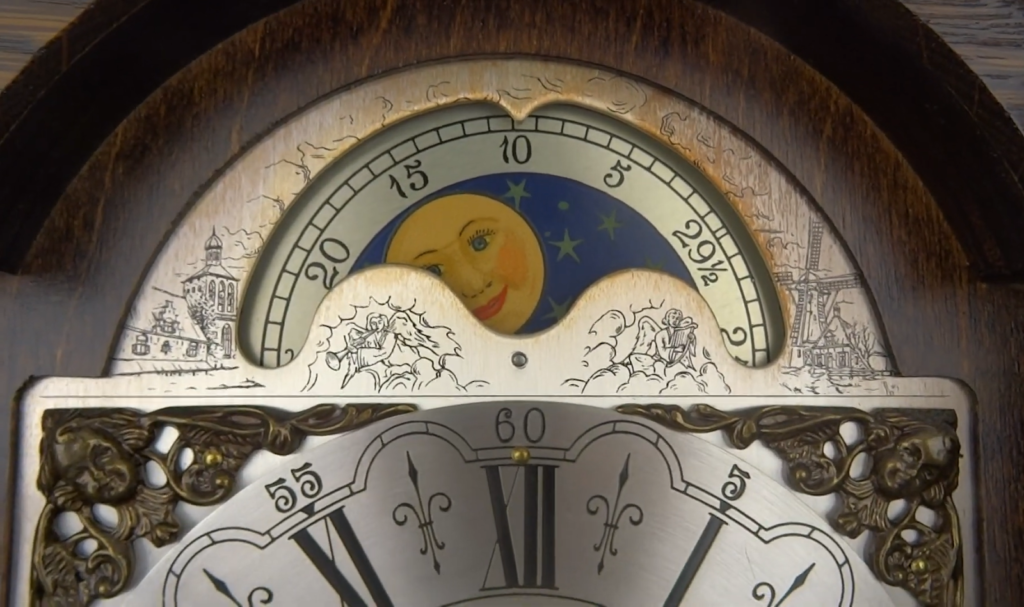 clock with golden moon with a smiling face against a blue background adorned with stars, positioned over detailed engravings and numbers